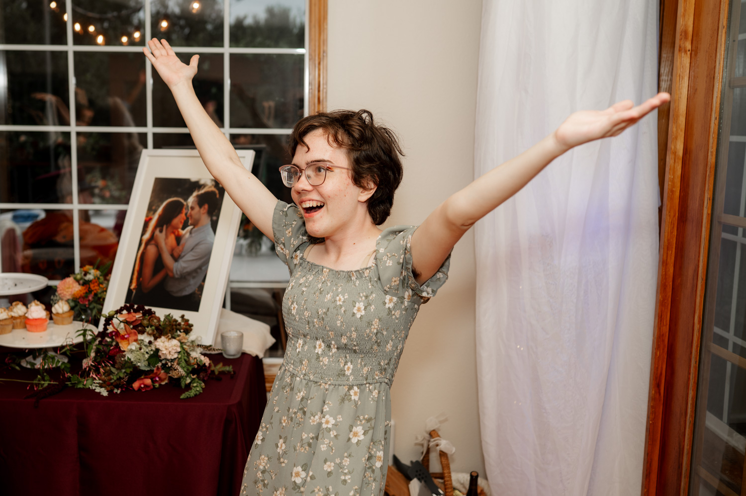 A joyful person in a floral dress raises their arms in celebration next to a table with wedding photos and vibrant flowers at the Yachats Inn.