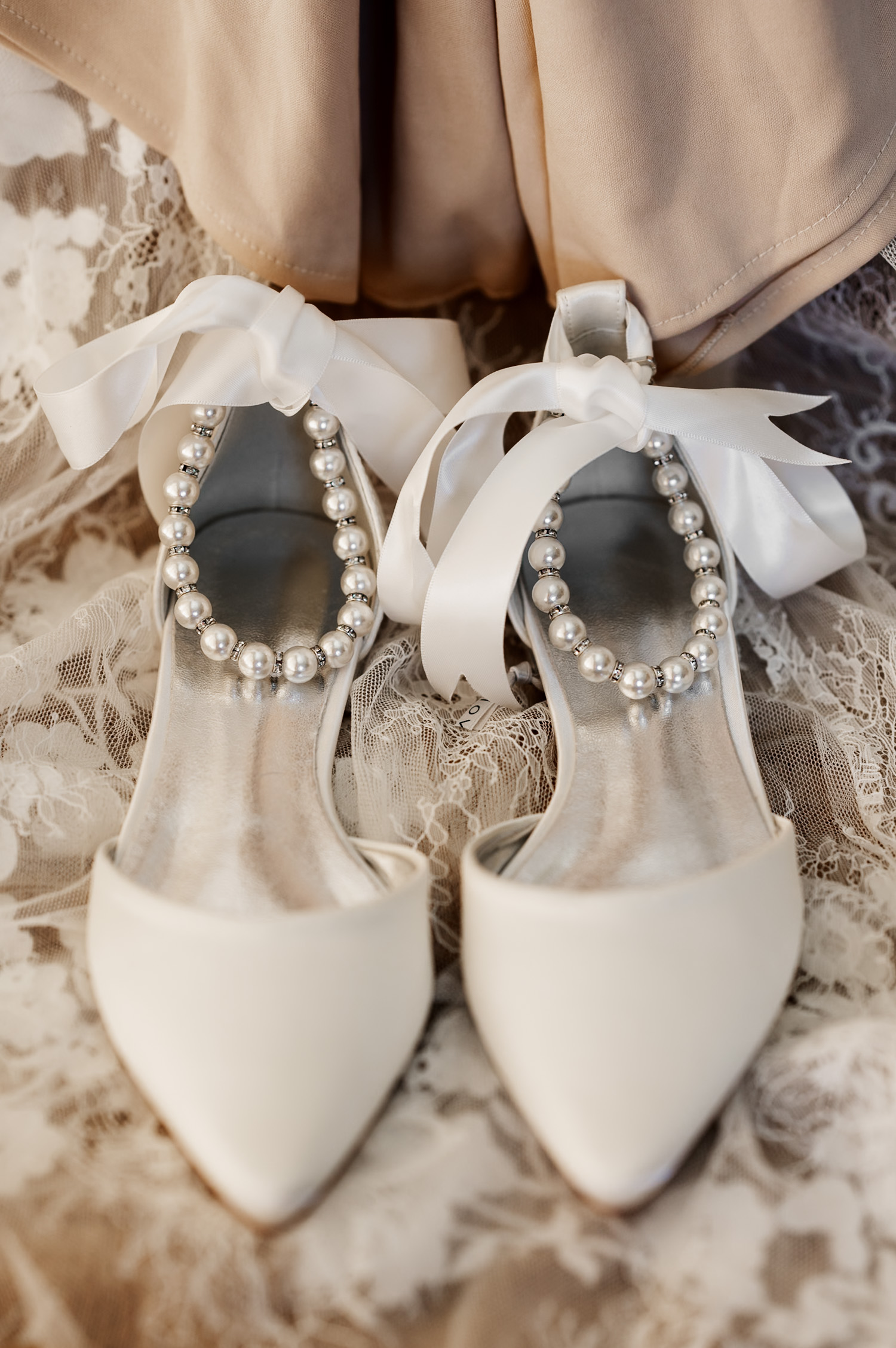 White bridal shoes featuring pearl ankle straps and ribbon bows, placed on lace fabric.