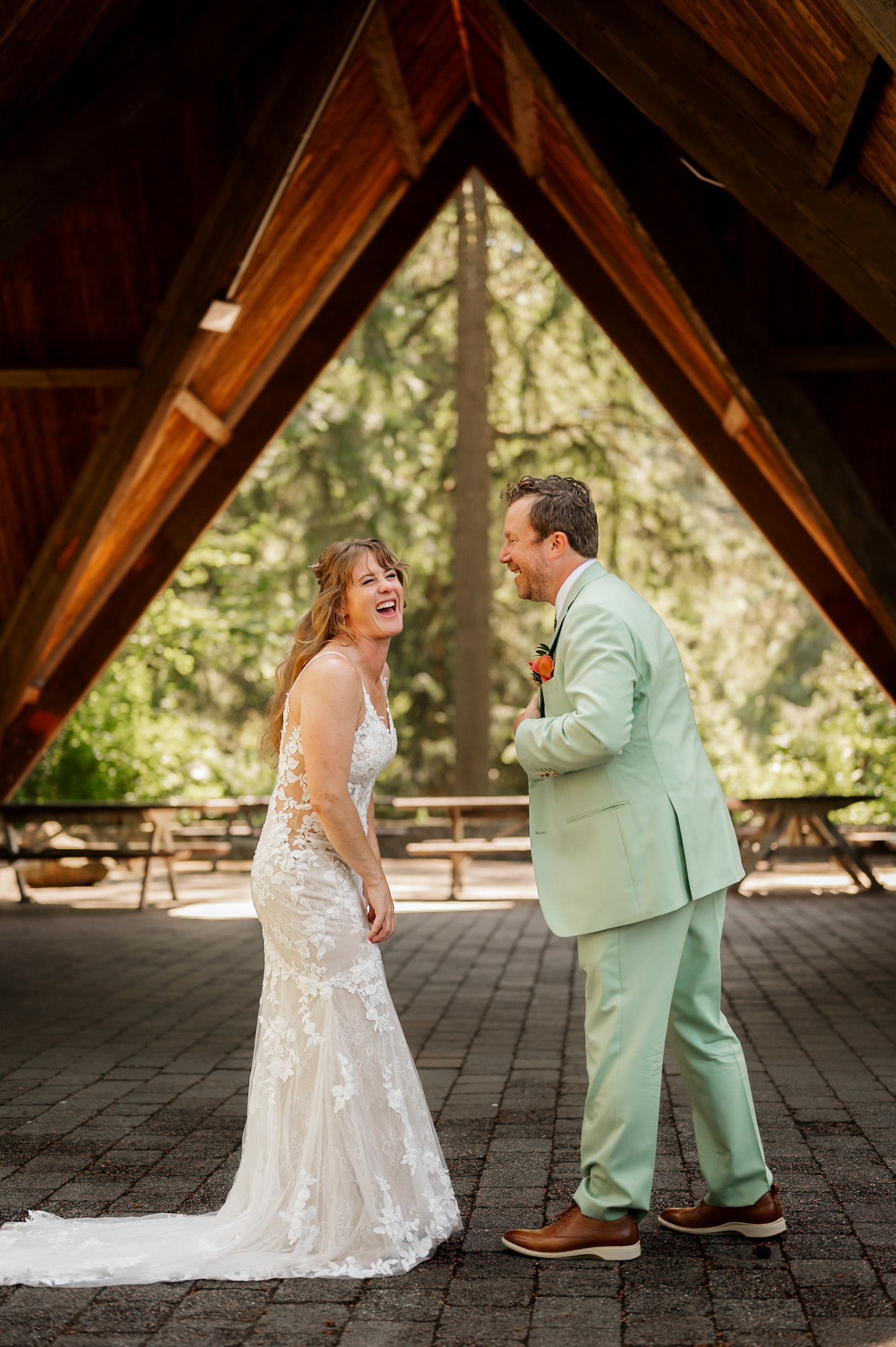 Bride in lace gown and groom in mint suit laughing together under a wooden structure at Hoyt Arboretum Stevens Pavilion.
