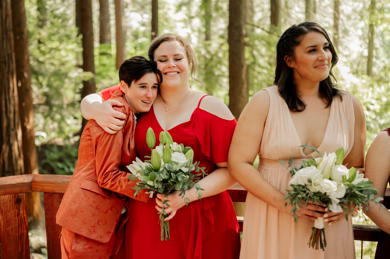Three people in colorful dresses, holding bouquets and smiling during wedding ceremony photos at the Redwood Deck in Hoyt Arboretum, with a lush forest backdrop.