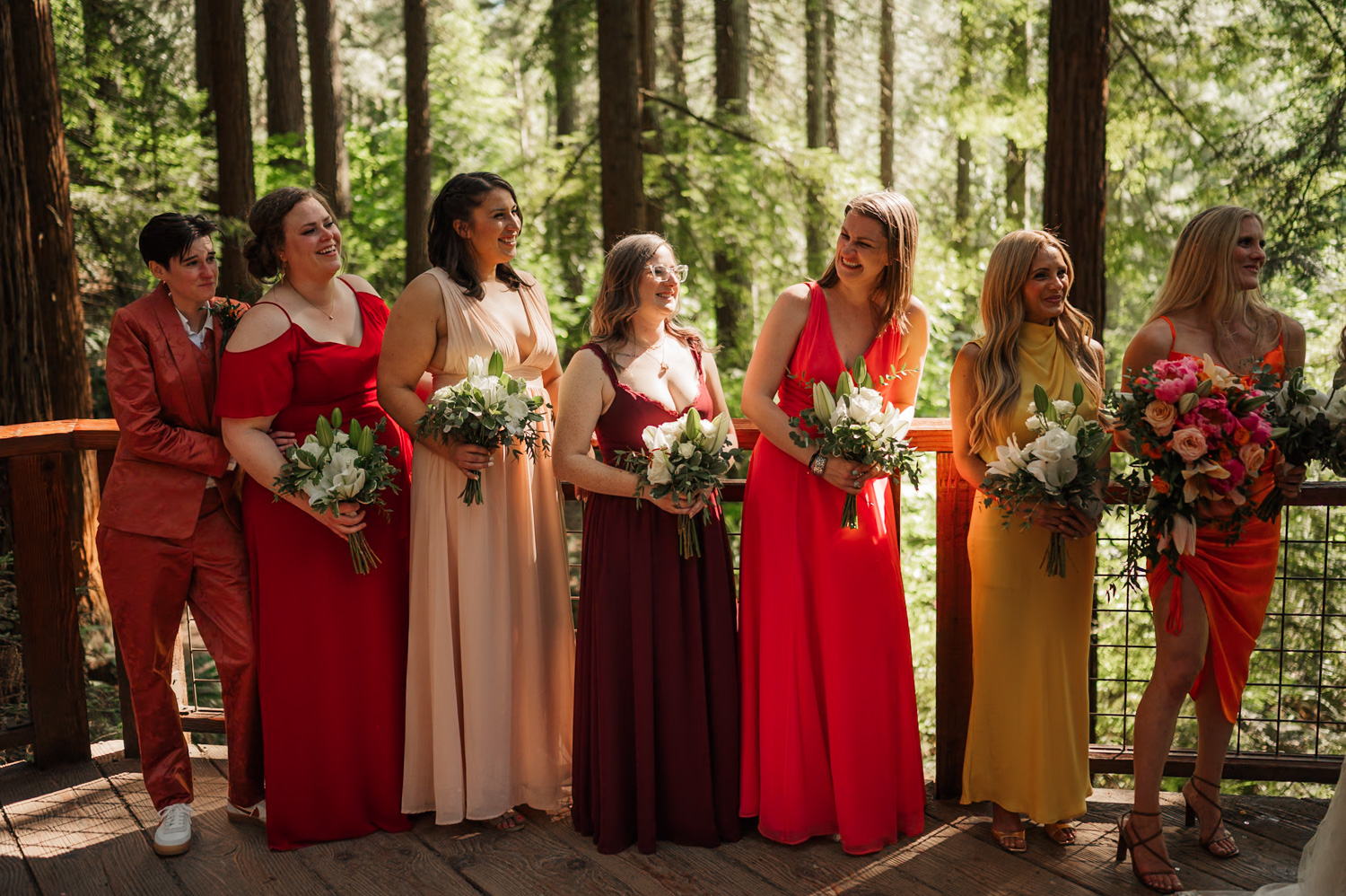 Seven people in colorful dresses and suits stand on a wooden deck surrounded by trees, holding bouquets. This photo was taken during a wedding ceremony at the Redwood Deck in Hoyt Arboretum.