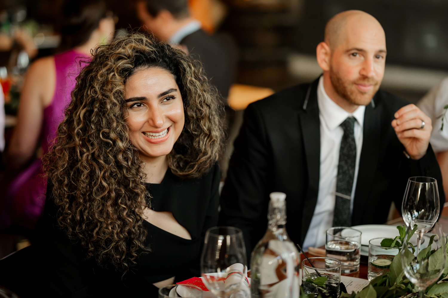 Two people sitting and smiling at a table with glasses and a bottle, enjoying the lively atmosphere of a wedding reception at Coopers Hall.