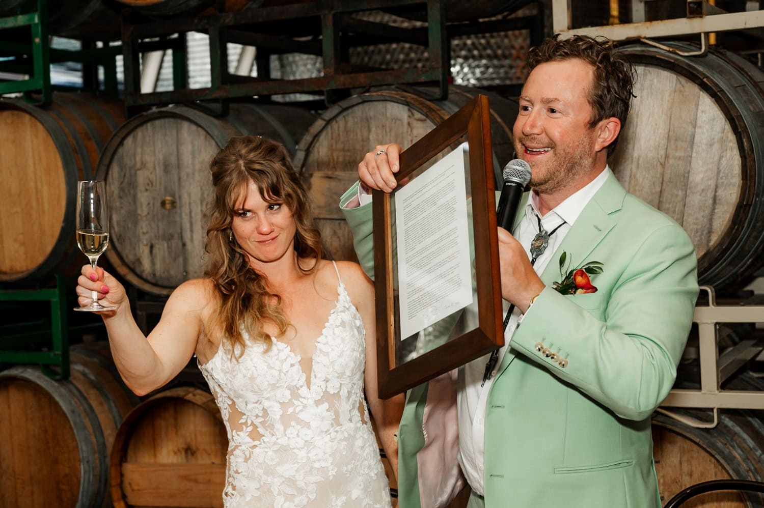 A bride holding a champagne glass stands next to a man in a mint suit at Coopers Hall. The man is holding both a framed document and a microphone during the wedding reception.