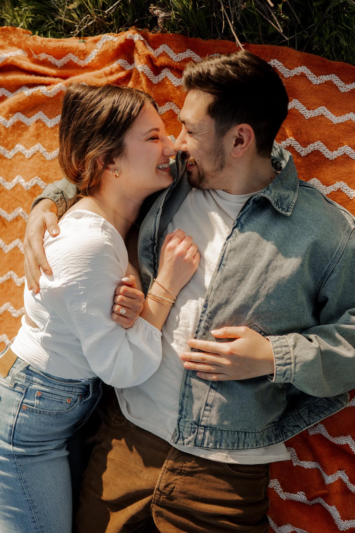 A couple laughs and cuddles together on an orange patterned blanket outdoors, surrounded by wildflowers at Rowena Crest during their engagement session.