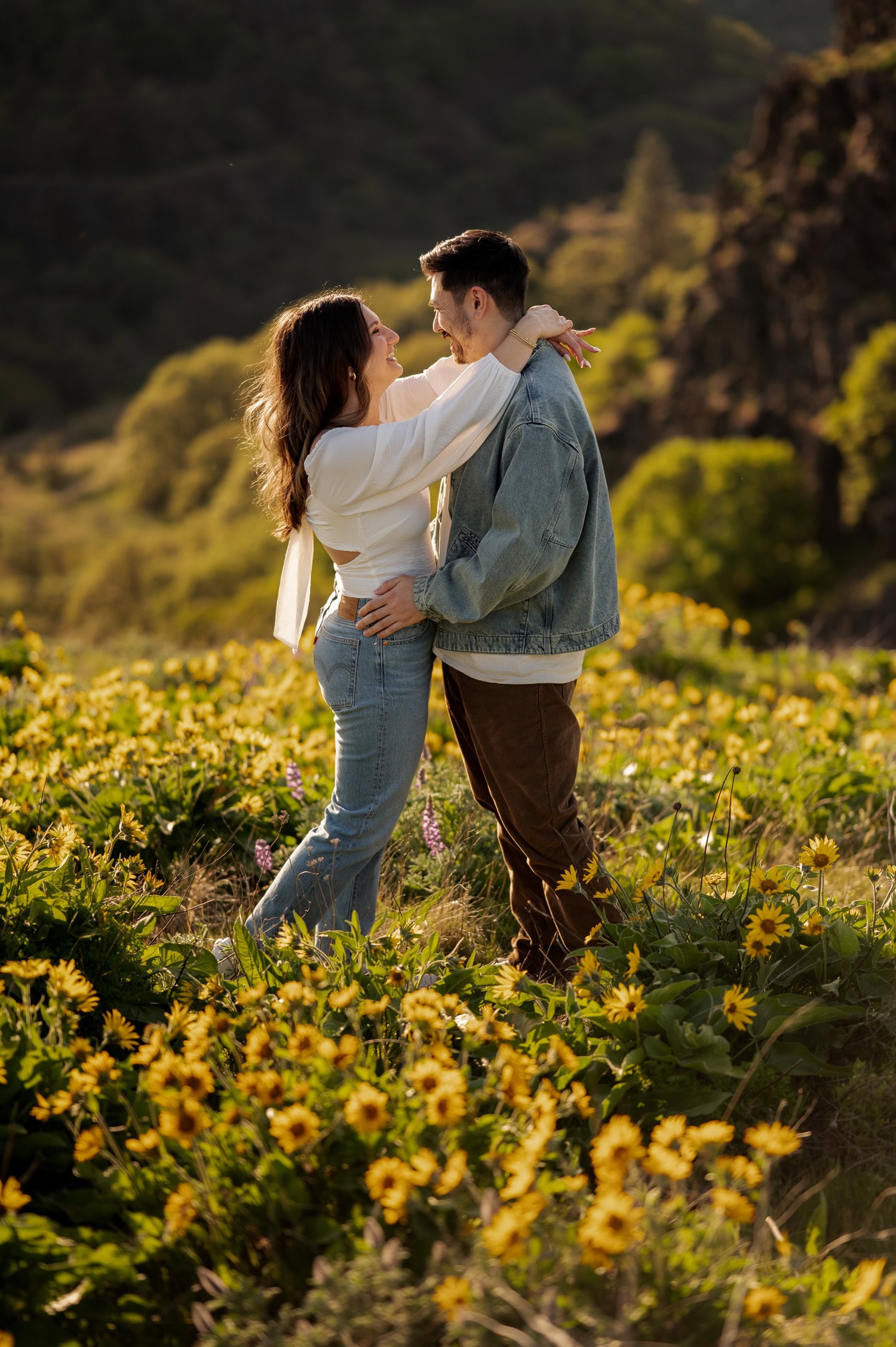 A couple embraces and smiles at each other in a field of wildflowers, with rolling green hills and Rowena Crest visible in the background.