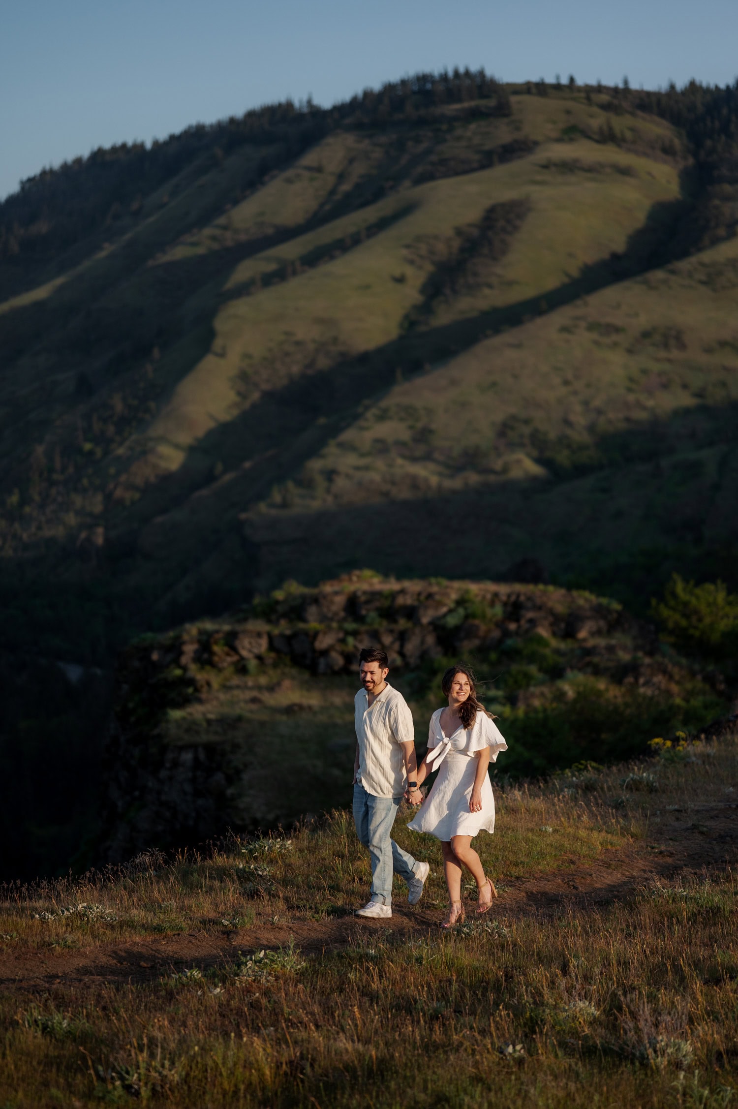 A couple holding hands walks along a grassy trail at Rowena Crest, surrounded by blooming wildflowers and rolling green hills under a bright sky.