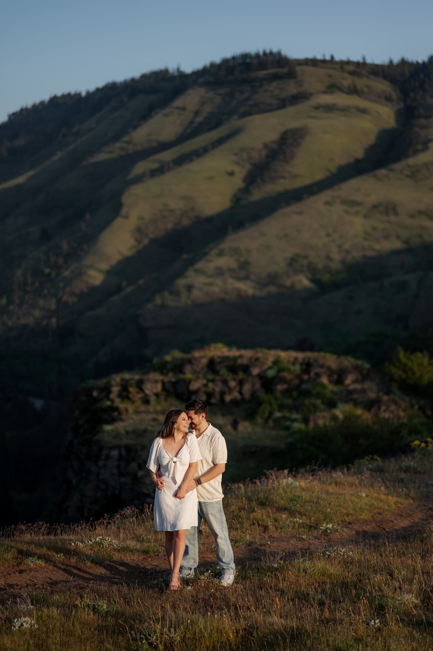 A couple stands close together on a grassy hill at Rowena Crest, surrounded by wildflowers with sunlit mountains in the background.