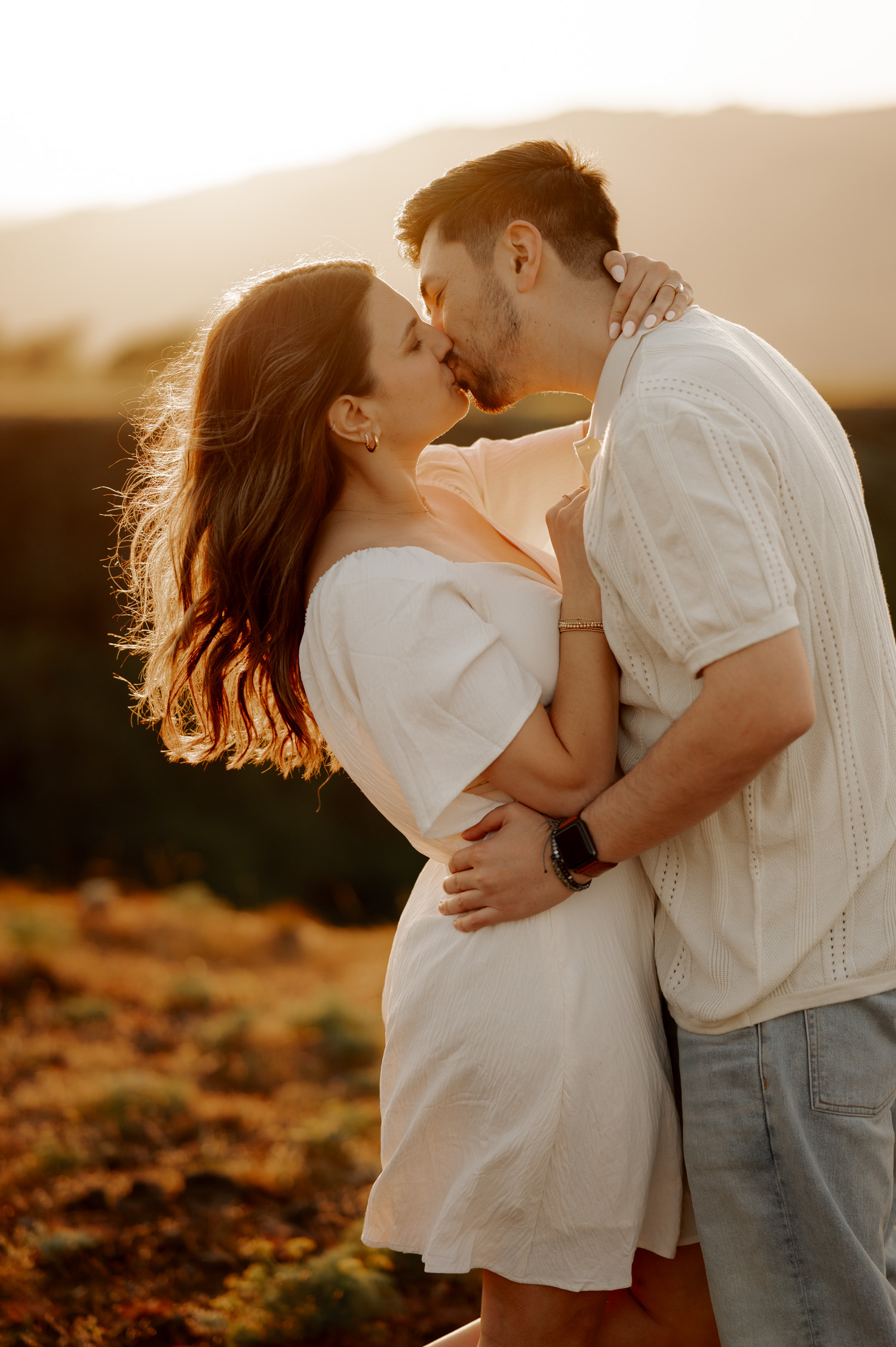 A couple in white outfits kisses at sunset on Rowena Crest, surrounded by wildflowers and illuminated by soft sunlight.