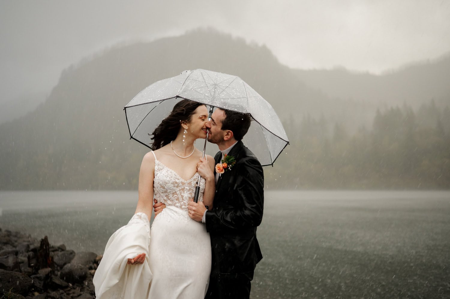 Bride and groom standing under a clear umbrella beside the Columbia River, with mountains in the background on their elopement day.