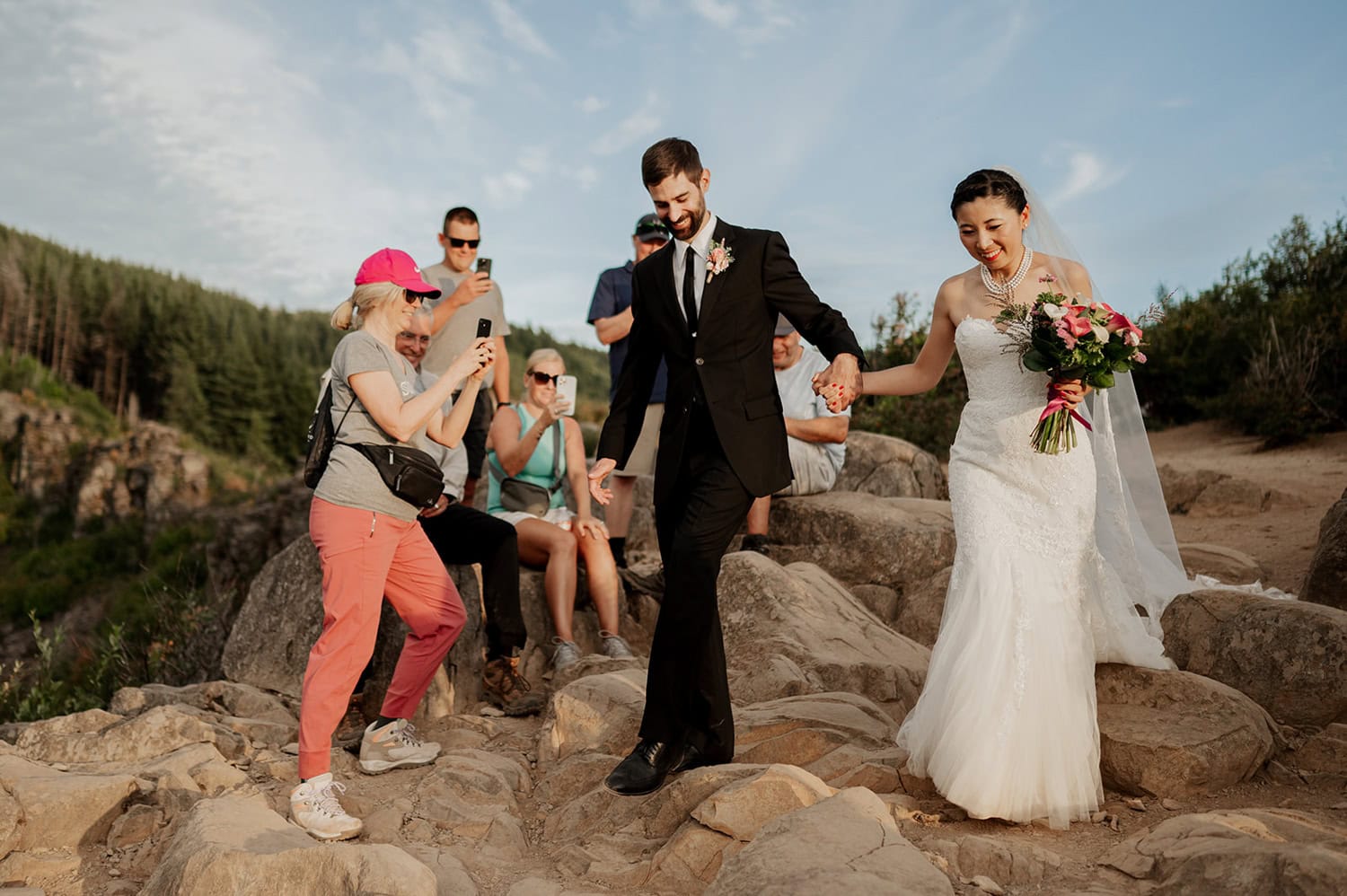 Bride and groom walk along a rocky path at Angel's Rest in the Columbia River Gorge while guests take photos, surrounded by nature, highlighting an outdoor elopement ceremony.
