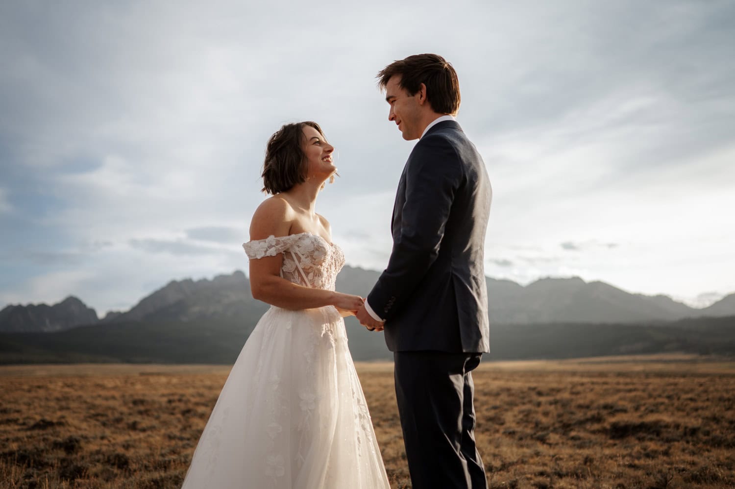 Bride and groom holding hands, smiling together at sunset in a field at the base of Stanley, Idaho's Sawtooth mountains.