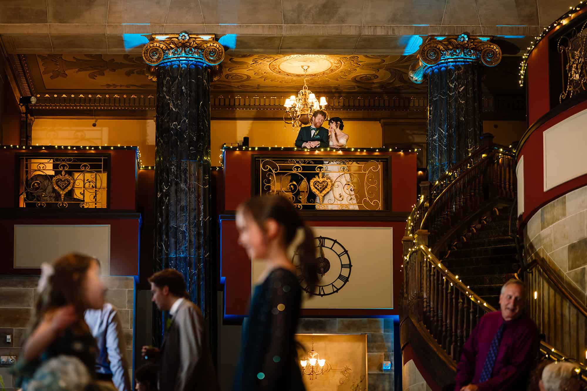 Emma and Terrance pose on The Society Room's upper balcony beneath a gilded chandelier