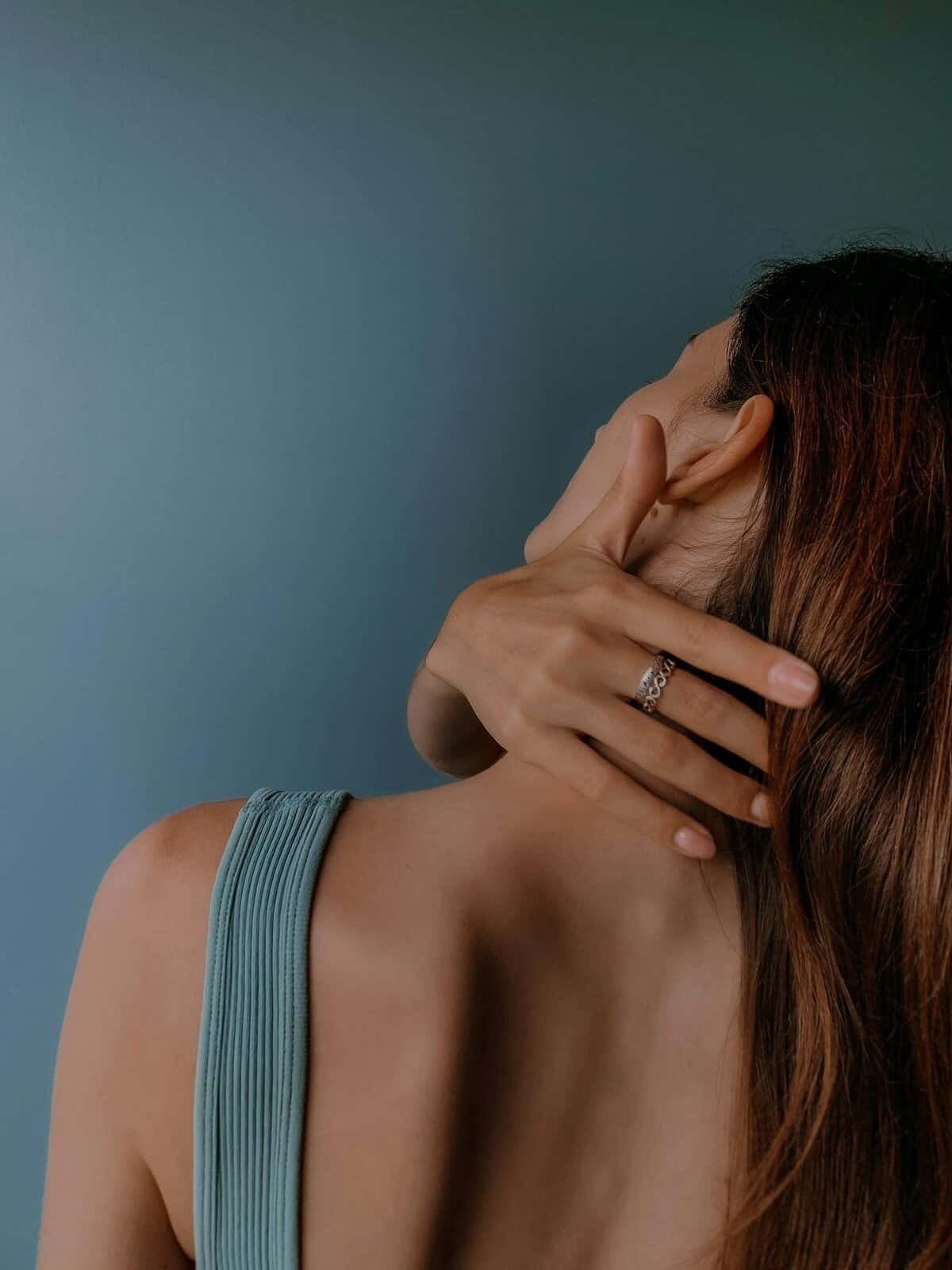 Close-up of a woman with long hair, hand on neck, wearing silver rings on a blue backdrop.