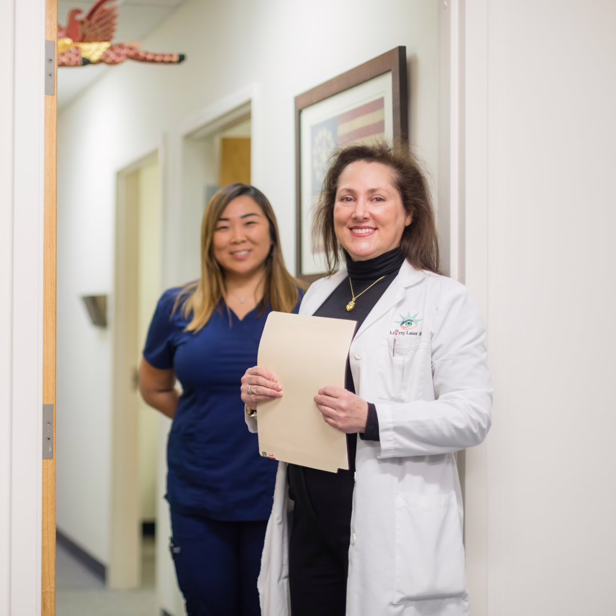 Two women, one in a lab coat and one in scrubs, stand in a hallway.