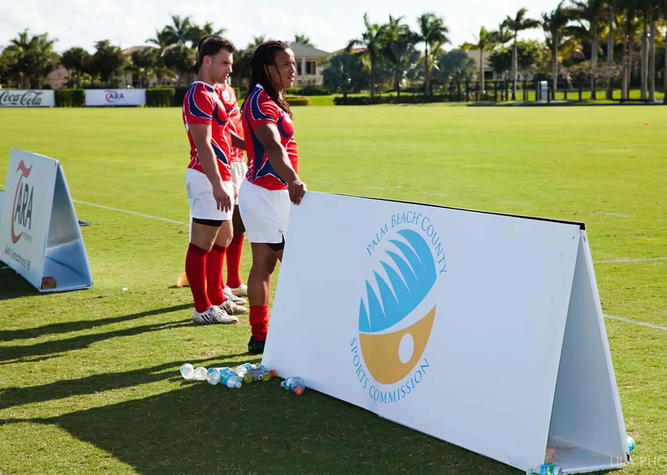 Two soccer players standing near a sign at a sports field with palm trees in the background.