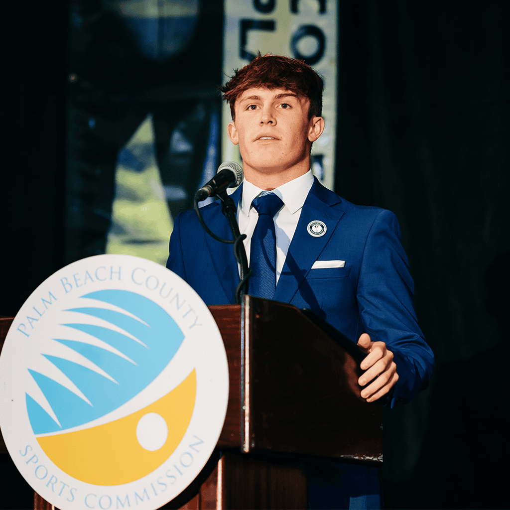 Young man in blue suit speaking at Palm Beach County Sports Commission event.