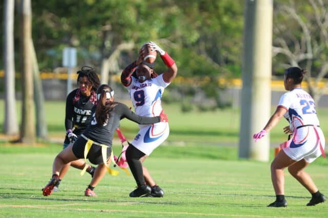 Female football player in white jersey catches ball during game with opponents.