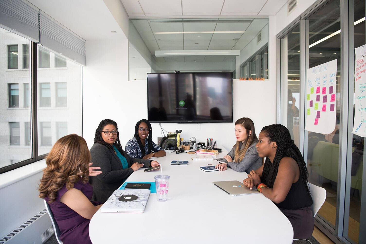 Several women sit around a conference table. A large digital display is mounted on the wall next to them available for people to join the meeting digitally.
