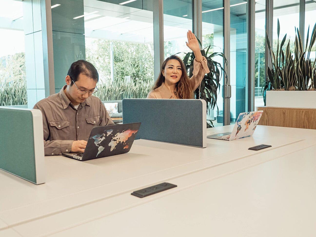 two people sitting at individual workstations, one looks at computer, the other looks at camera and is raising their hand
