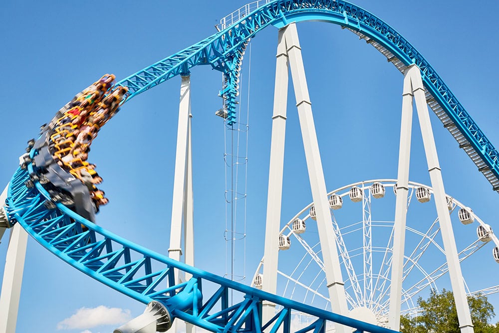 A blue roller coaster with a train of riders speeds down a curved track, showcasing the excitement of the Makeway Videri partnership, with a Ferris wheel and clear blue sky in the background.