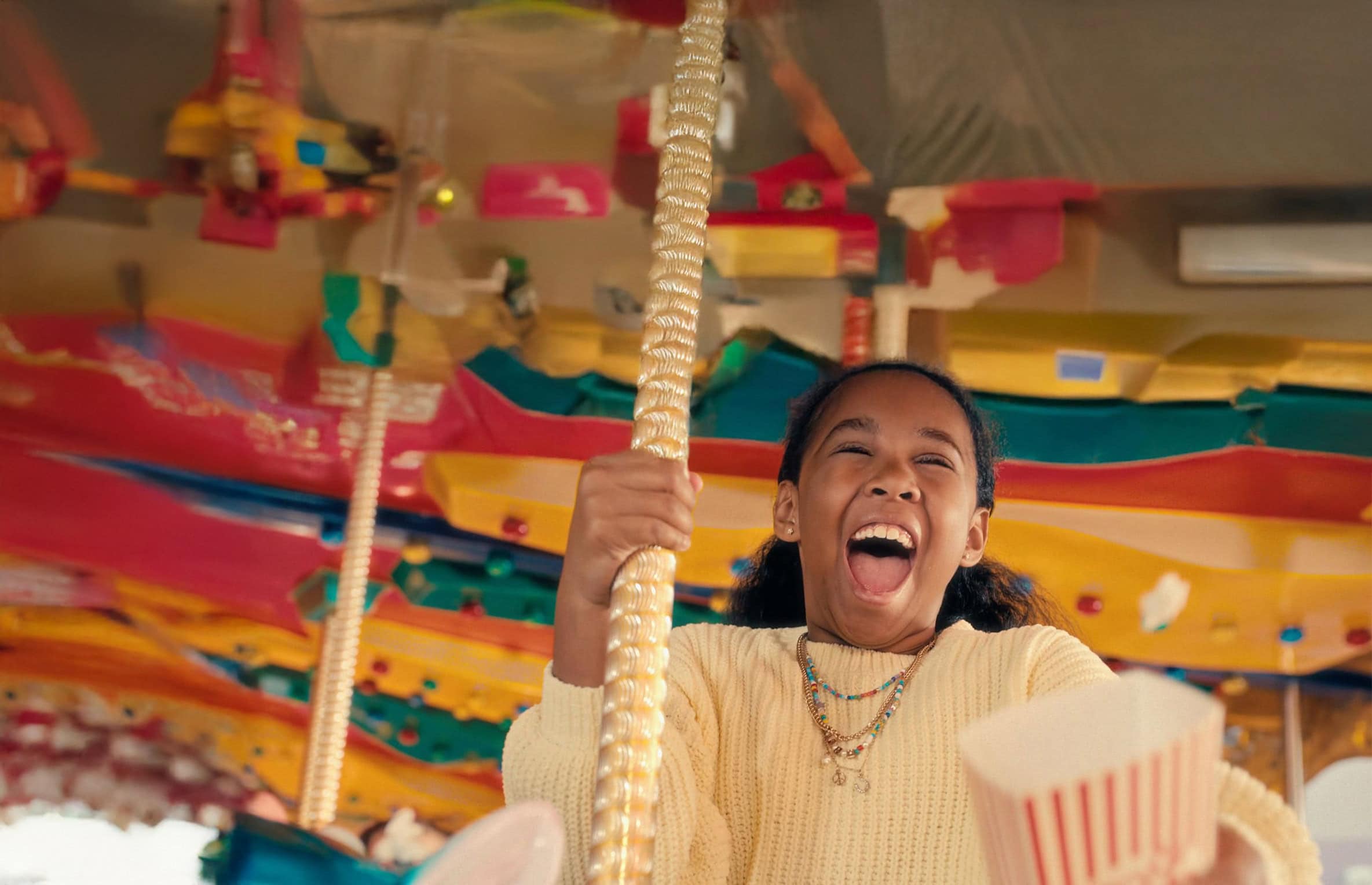 A girl in a cream sweater laughs while riding a colorful carousel, holding a pole, as a popcorn container marked with the Makeway Videri partnership peeks out in the foreground.