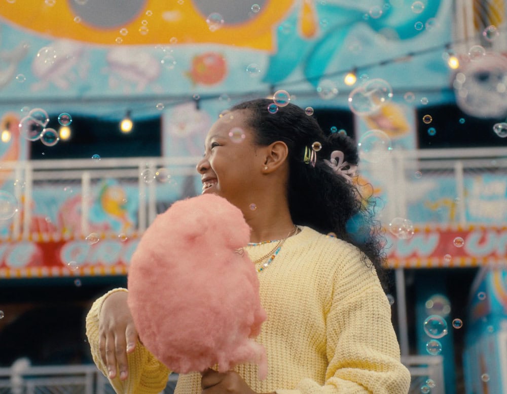 A girl in a yellow sweater holds pink cotton candy and smiles, surrounded by bubbles, with a colorful amusement park backdrop, celebrating the joy of the new Makeway Videri partnership.