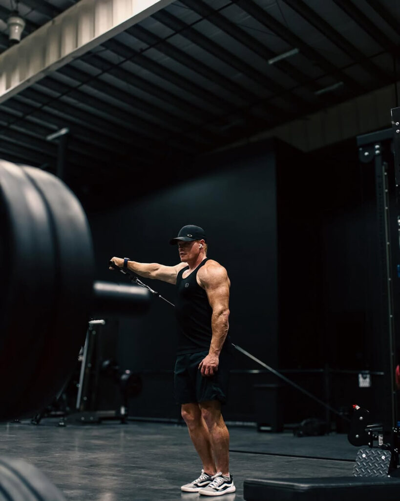 Brent in athletic wear performs a cable lateral raise exercise in a dimly lit gym, holding the handle with his right hand at shoulder height.