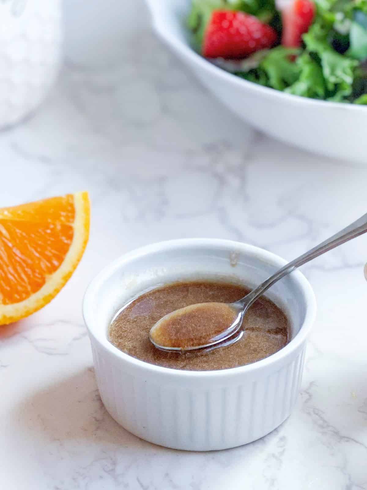 Balsamic citrus vinaigrette in a white ramekin with spoon, strawberry salad, and sliced oranges.