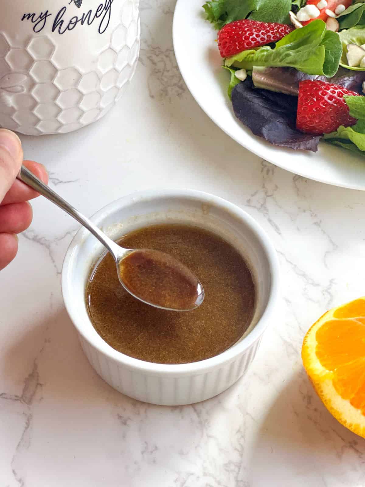 Balsamic citrus vinaigrette in a white ramekin with spoon, strawberry salad, honey jar, and sliced oranges.