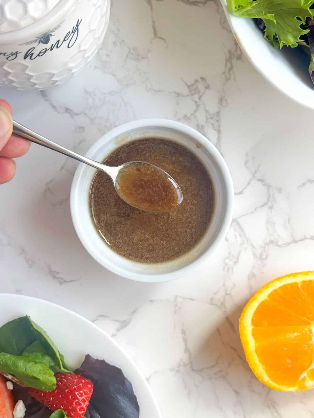 Balsamic citrus vinaigrette in a white ramekin with spoon, strawberry salad, honey jar, and sliced oranges.