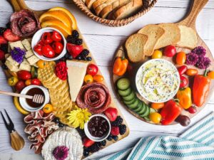 Wooden charcuterie boards with small dishes, cheeses, meats, veggies, fruits, and flowers.