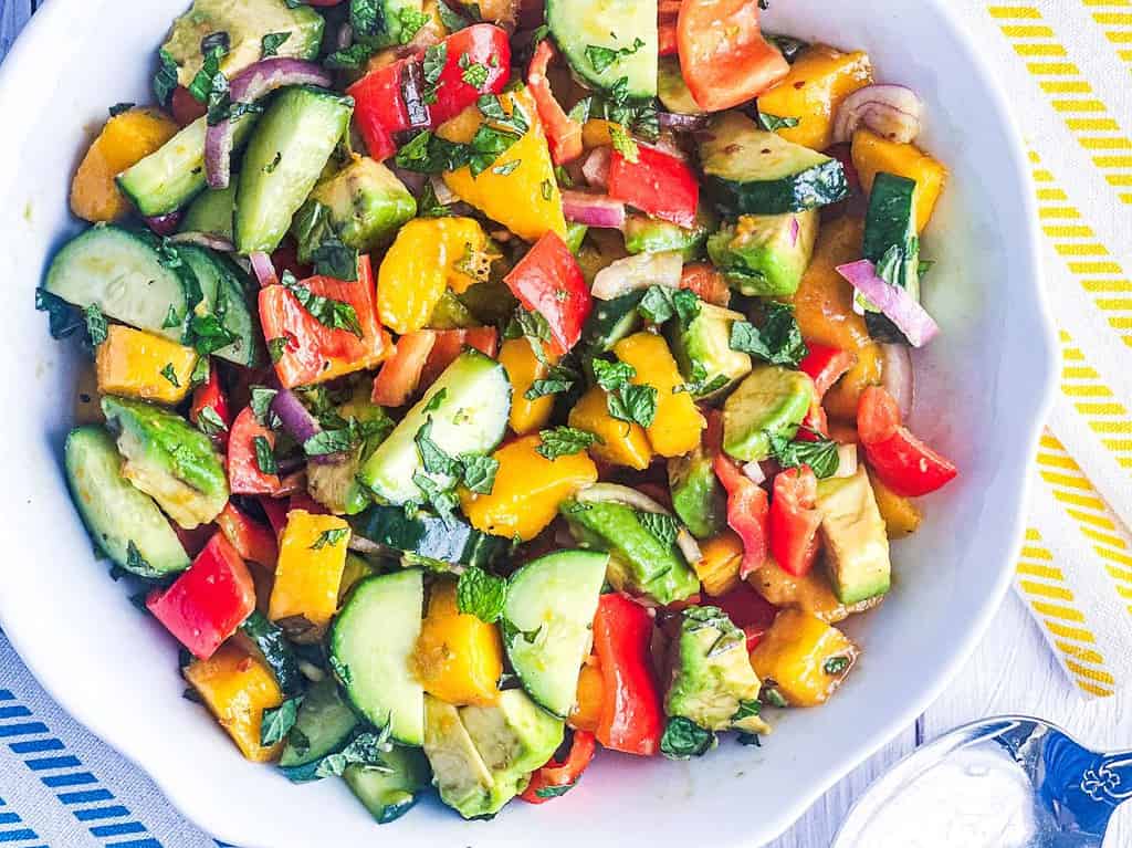 Avocado, cucumber, mango, and bell pepper salad with shallots and fresh mint in a white bowl with a spoon on the side.