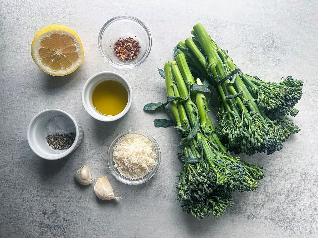 Broccolini, lemon, red pepper flakes, parmesan, garlic, olive oil, salt, and pepper on gray background.