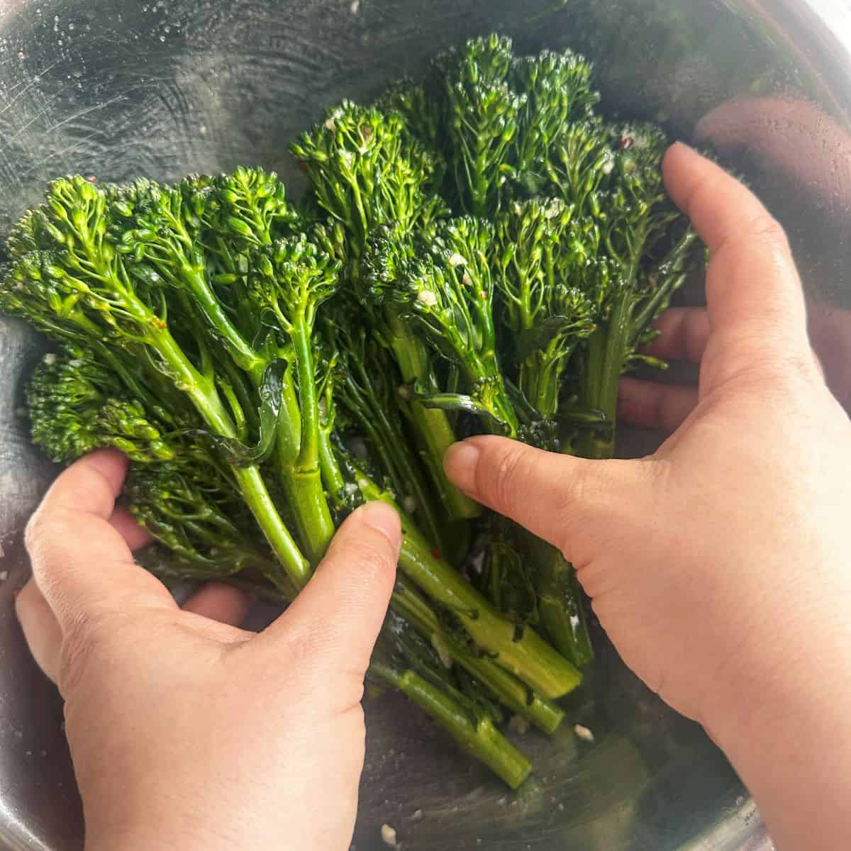 Broccolini in a bowl with hands mixing the dressing.
