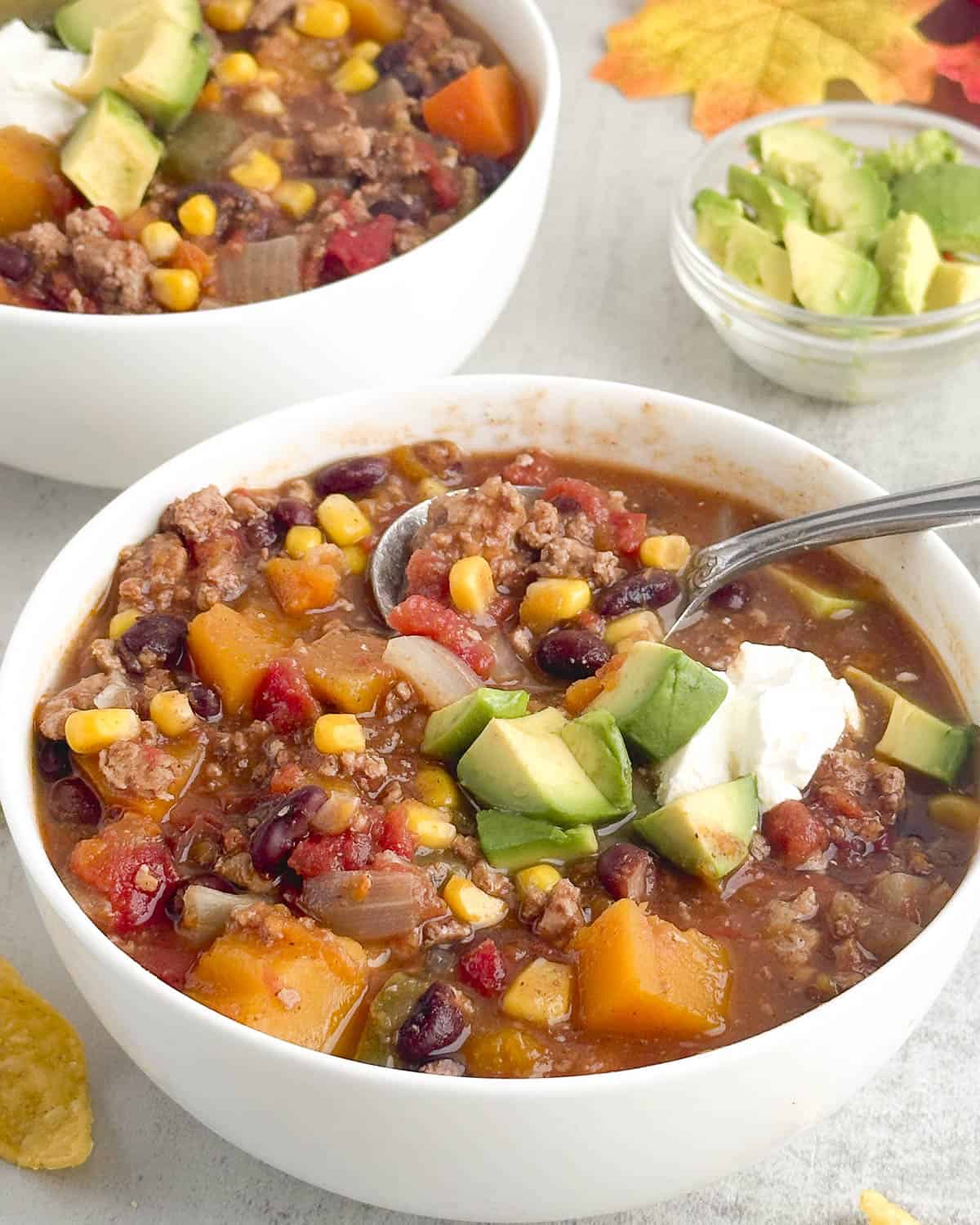 Fall harvest chili with avocado and sour cream garnish in a white bowl with a spoon dipping in.