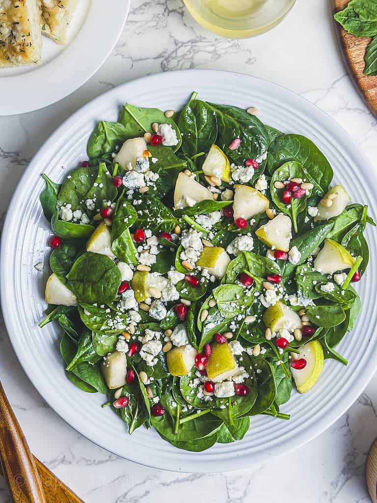 Spinach, Gorgonzola Cheese, Pear, Pomegranate, and Pine Nuts in a white serving bowl.