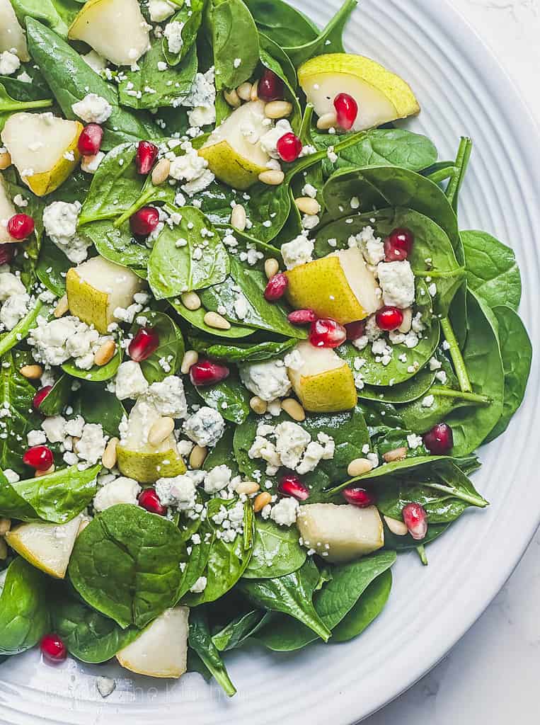 Spinach salad with blue cheese, pears, pomegranate, and pine nuts in a white serving bowl.