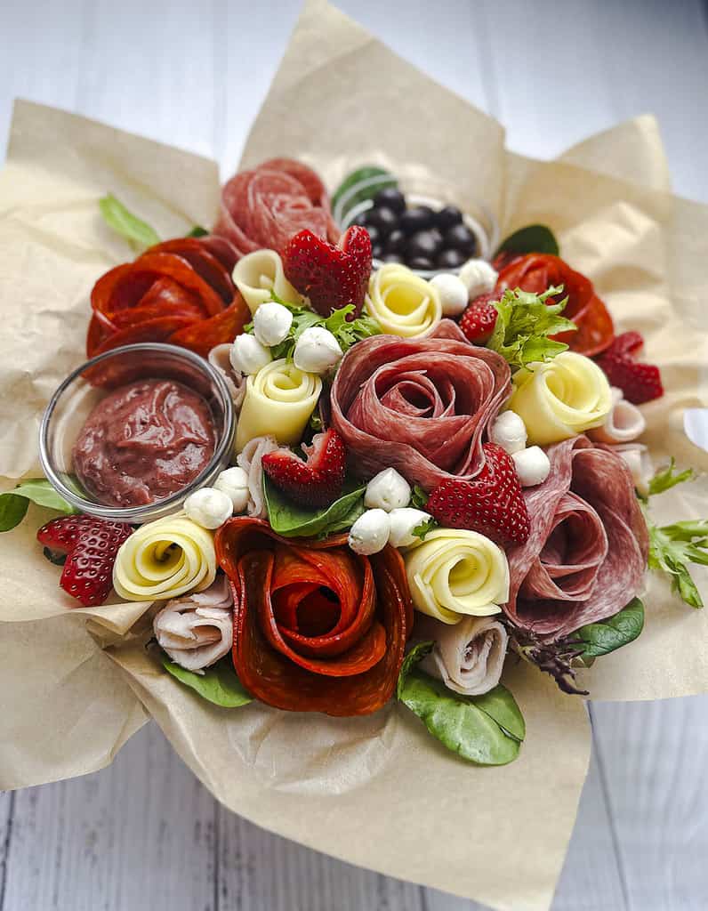 A charcuterie bouquet with meat and cheese roses with fresh greens, mini mozzarella balls, and heart-shaped strawberries.