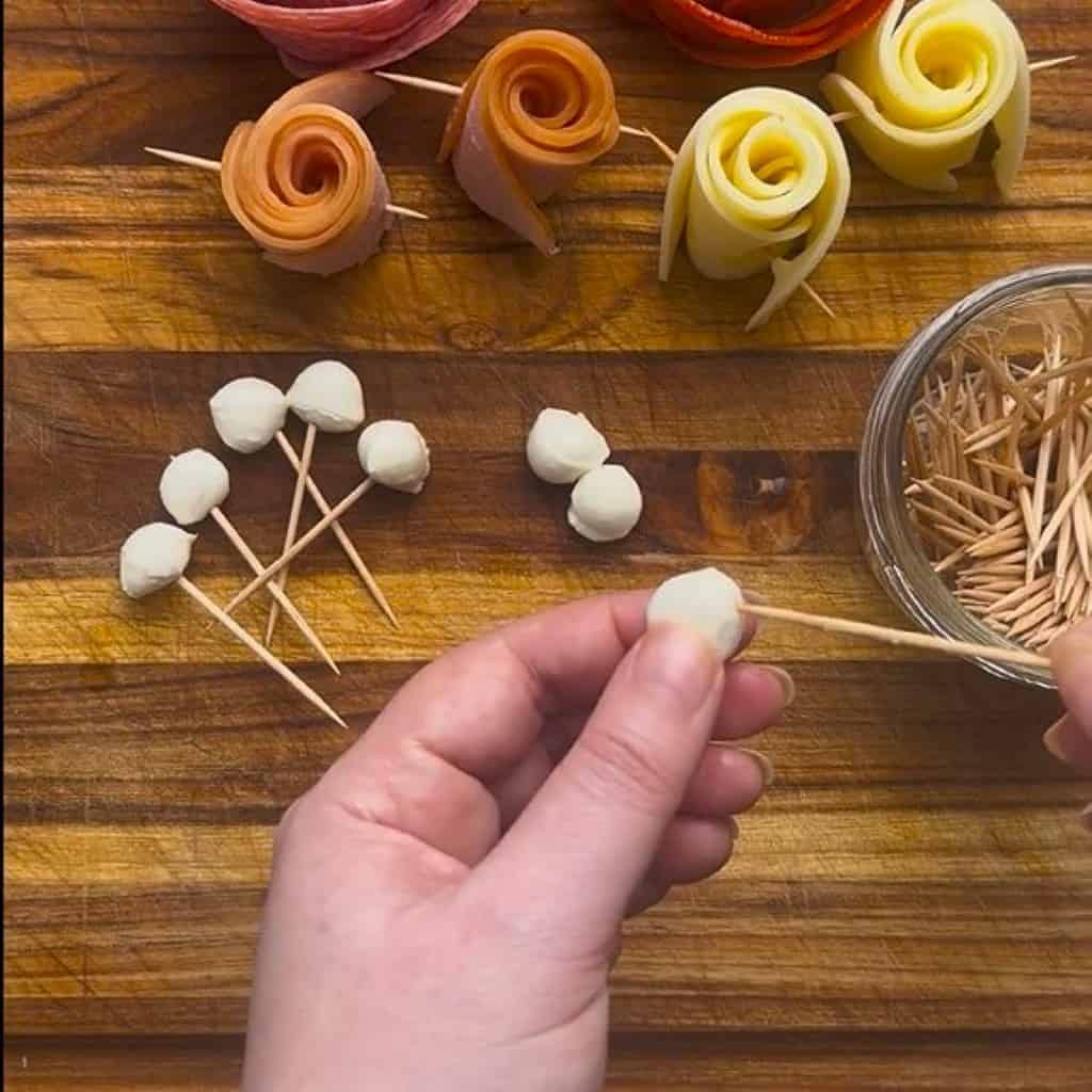 Process shot of hands adding a mozzarella pearl to the end of a toothpick.