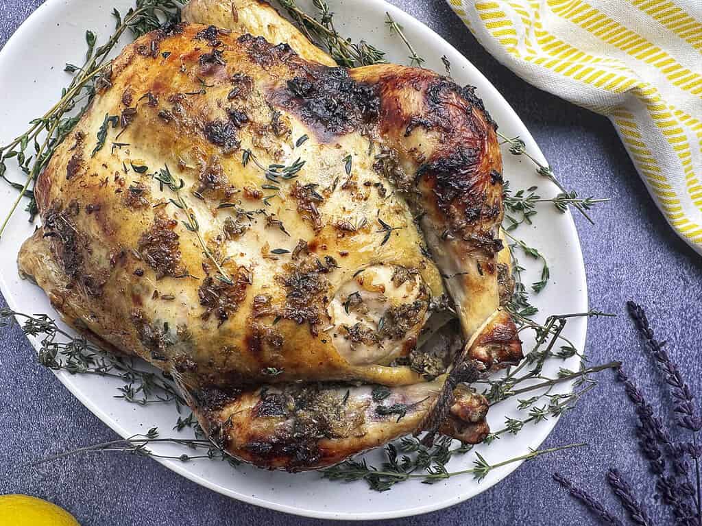 Whole roasted chicken, trussed and displayed on a white oval serving platter surrounded by fresh thyme springs and lavender.