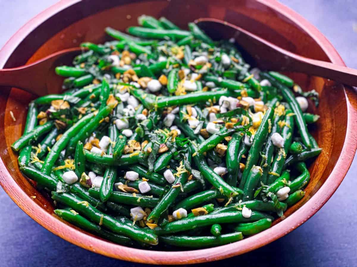 Green beans in a wooden bowl topped with goat cheese and pistachios and with two serving spoons.