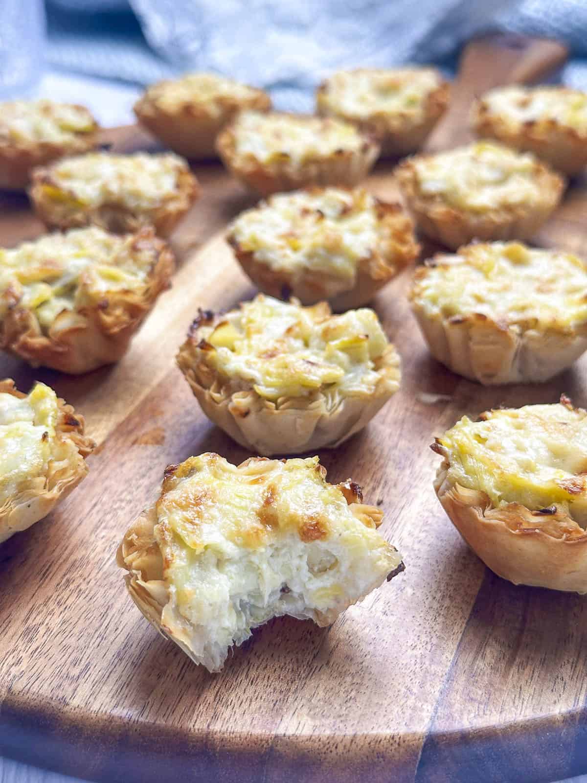 Artichoke tartlets on a wooden board with a bite missing from the one in front.