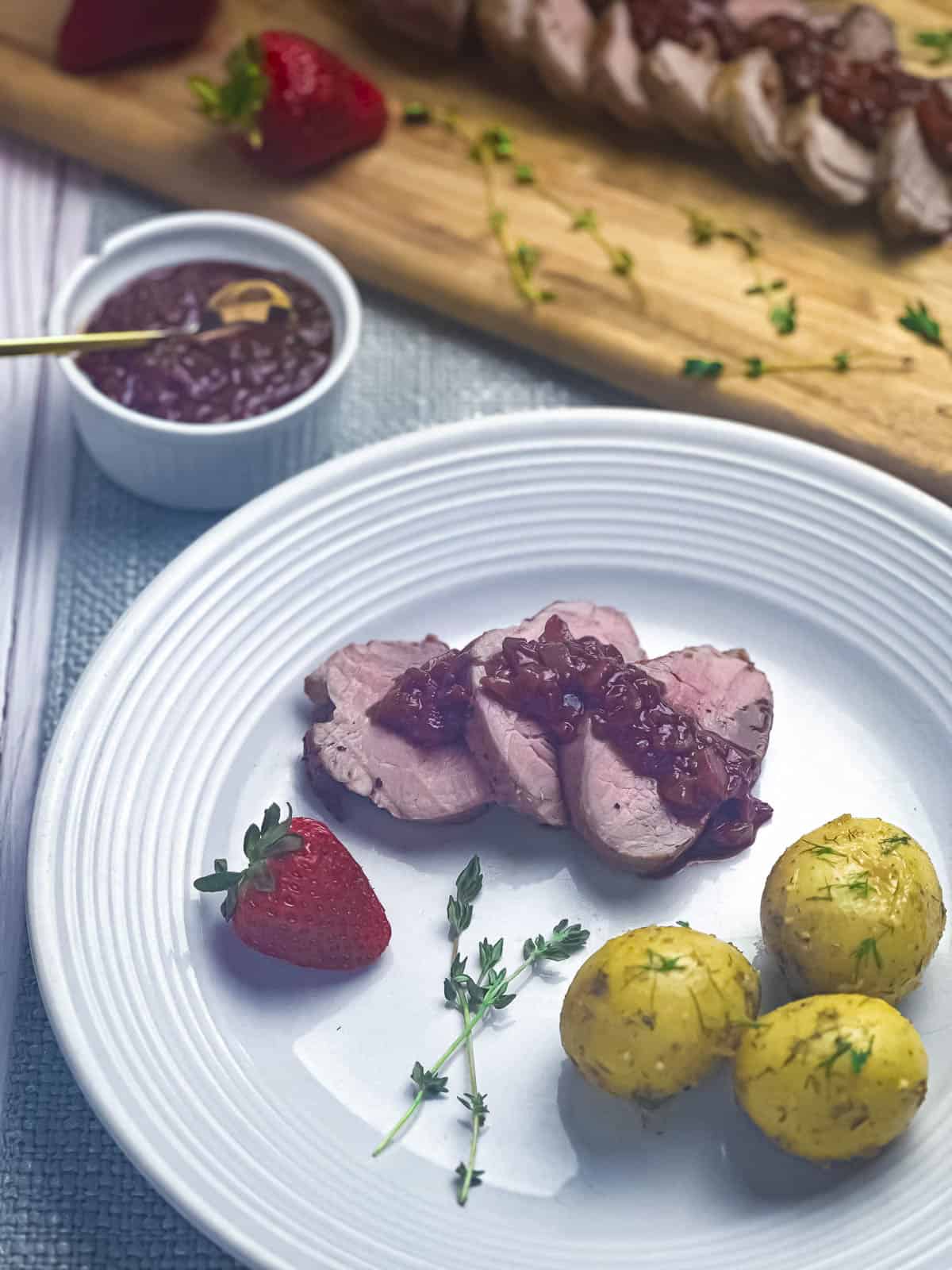 Slices of pork tenderloin with balsamic strawberry sauce, new potatoes, sprigs of thyme and a strawberry on a white dinner plate.