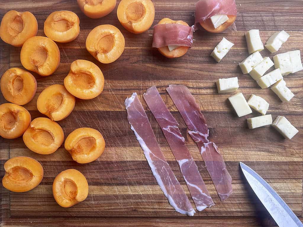 Strips of prosciutto, halved apricots, cheese cubes, with knife on a wooden cutting board.