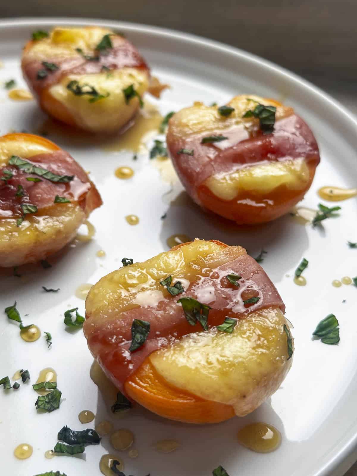 Halved apricots topped with melted Brie cheese, wrapped with prosciutto, and drizzled with honey on a white plate.