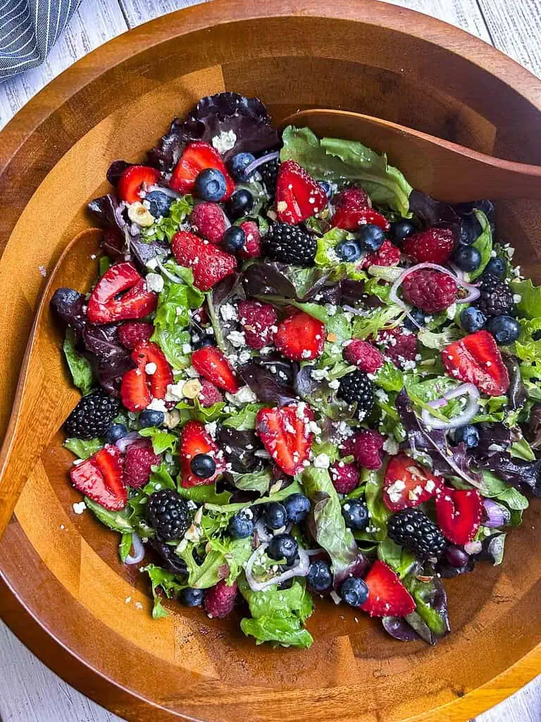 Berries, shallots, goat cheese crumbles, and mixed greens in a wooden salad bowl with a pair of serving spoons.