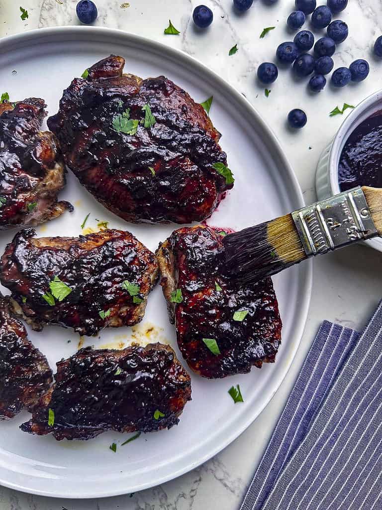 Barbecued chicken thighs on a plate being brushed with blueberry BBQ sauce.