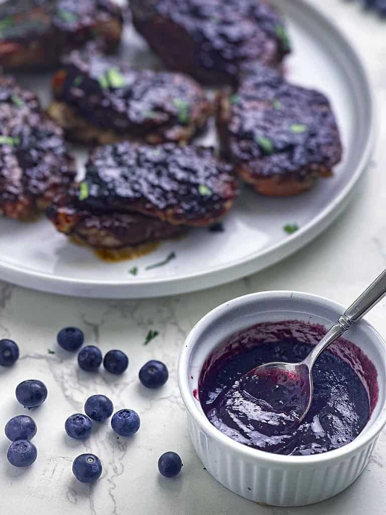 Blueberry BBQ sauce in a ramekin with a plate of barbecued chicken in the background.
