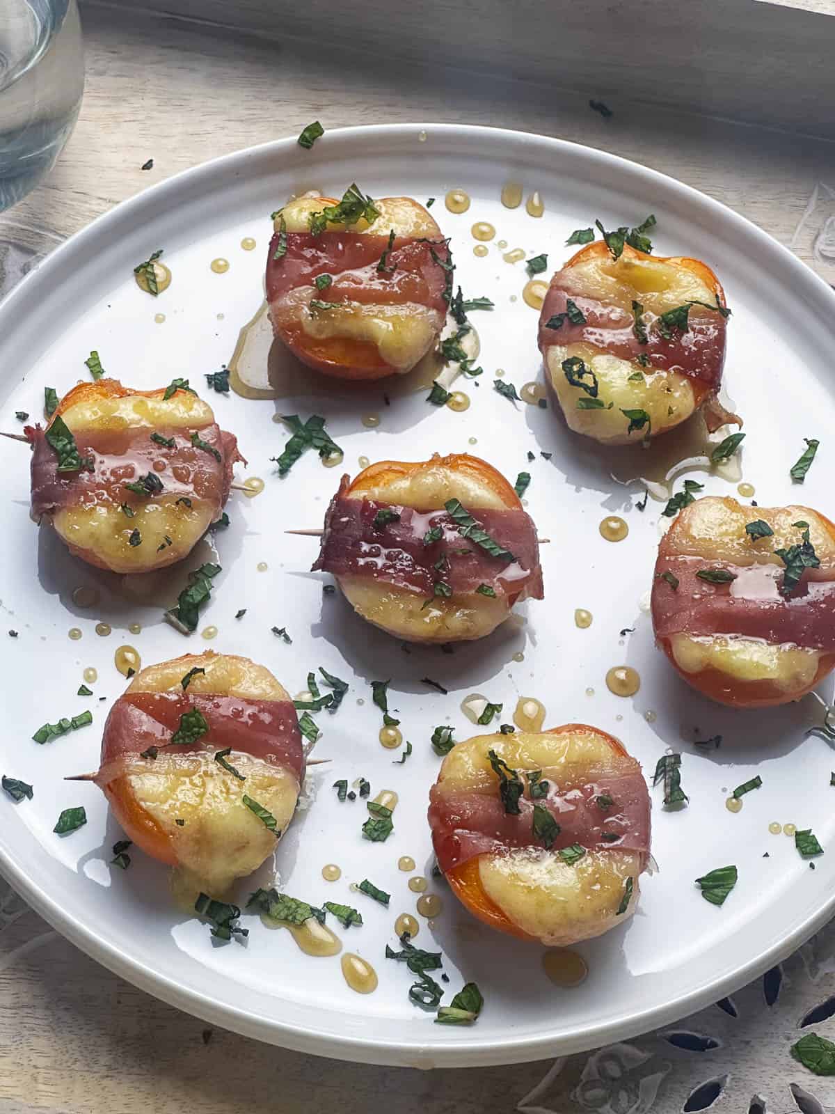 A white plate with apricot halves wrapped in prosciutto with melted Brie cheese and a drizzle of honey and chopped mint.