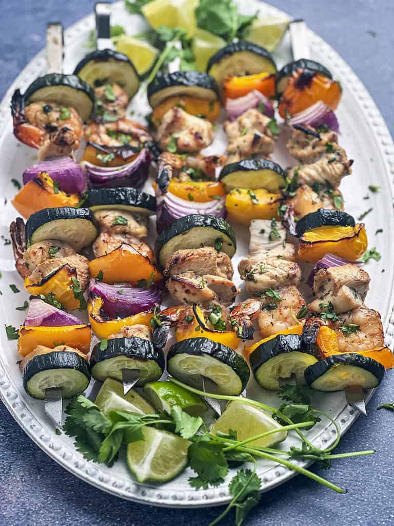 Grilled shrimp and chicken skewers with veggies on a white oval plate lime wedges and cilantro.